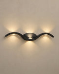 SCI Lighting Tidal black wave wall sconce lit with warm 3000K glow on a greige wall