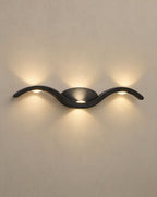 SCI Lighting Tidal black wave wall sconce lit with warm 3000K glow on a greige wall